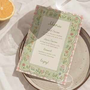 May include: A menu card with a floral border and a pink gingham background. The menu lists a first course of zucchini flowers, a main course of grilled duck breast, and a dessert of raspberry sorbet. The word "Menu" is at the top.
