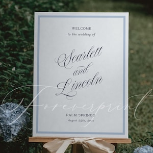 Könnte beinhalten: Ein Hochzeits-Willkommensschild auf einer Holzstaffelei. Das Schild hat einen hellblauen Rahmen und zeigt die Namen "Scarlett and Lincoln" in eleganter Schrift. Der Text "WELCOME to the wedding of" steht über den Namen, und "PALM SPRINGS August 25th, 20xx" steht darunter.