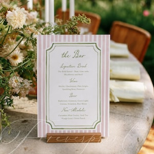 May include: A pink and white striped bar menu with a wooden base. The menu lists signature drinks, wines, beers, and non-alcoholic beverages. The text is in a decorative font with a green border.