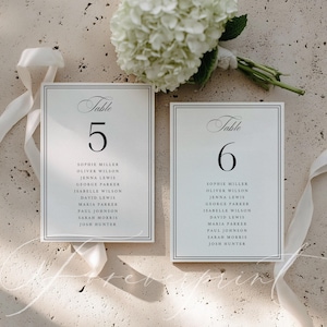 Puede incluir: Dos tarjetas blancas para números de mesa de boda con texto negro, con los números 5 y 6, y los nombres de los invitados. Las tarjetas están enmarcadas con un borde negro fino. Un ramo de flores blancas y una cinta blanca están en el fondo.