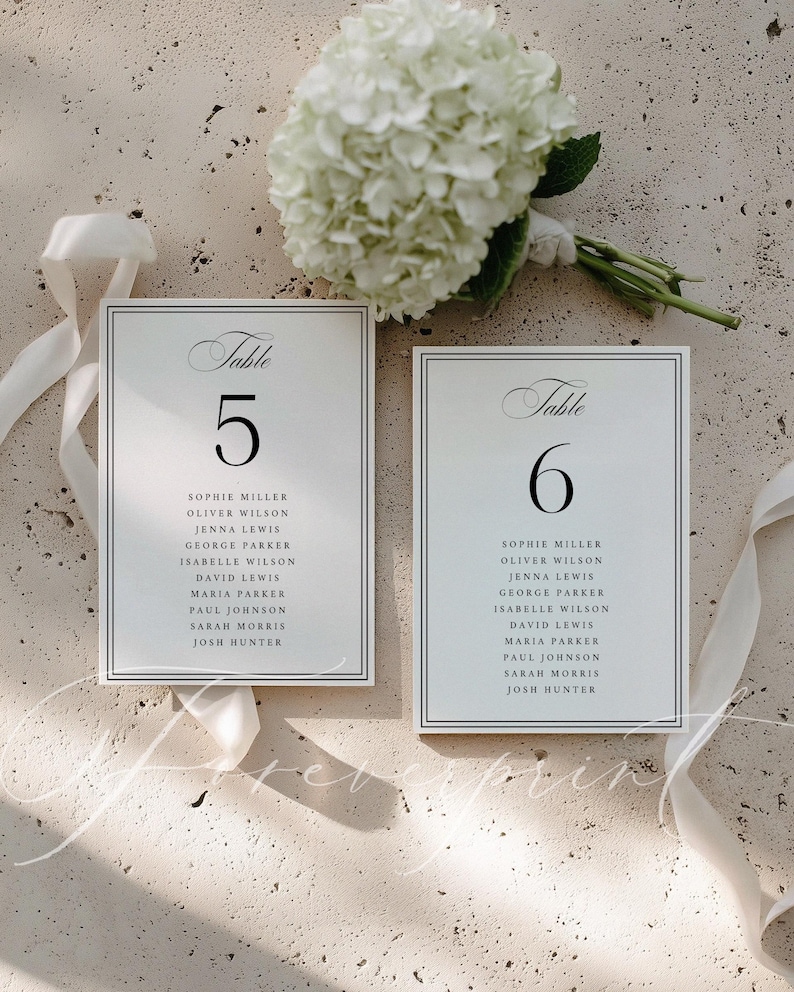 May include: Two white wedding table number cards with black text, featuring the numbers 5 and 6, and guest names. The cards are framed with a thin black border. A bouquet of white flowers and white ribbon are in the background.