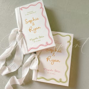May include: Two white wedding programmes with gold text reading Sophie & Ryan, Magnolia House, and 09.14.20xx. One programme features a pink wavy border and the other a green wavy border, both tied with long white silk ribbons on the left side.