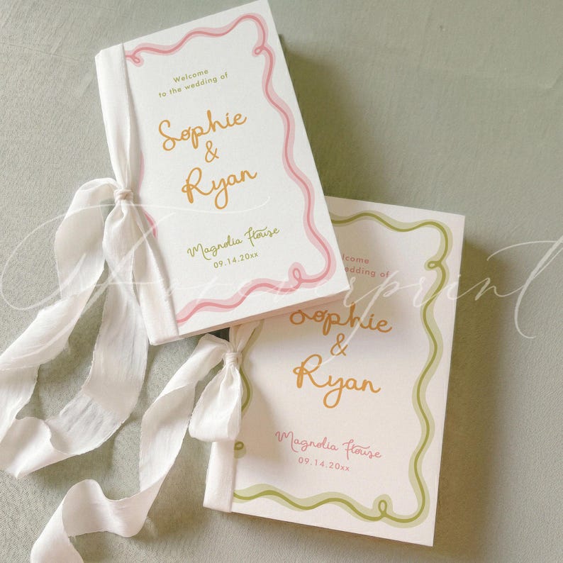 May include: Two white wedding programmes with gold text reading Sophie & Ryan, Magnolia House, and 09.14.20xx. One programme features a pink wavy border and the other a green wavy border, both tied with long white silk ribbons on the left side.