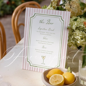 May include: A bar menu sign with a pink and white striped background. The sign features the text "the Bar" and a list of signature drinks, wine, and beer options. A small bowl of lemons sits in front of the sign, with a cocktail glass illustration.