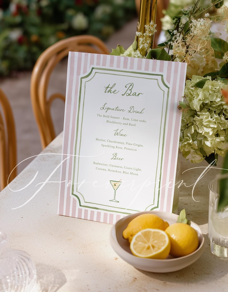 May include: A bar menu sign with a pink and white striped background. The sign features the text "the Bar" and a list of signature drinks, wine, and beer options. A small bowl of lemons sits in front of the sign, with a cocktail glass illustration.