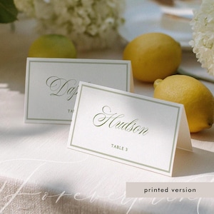 May include: Two white folded place cards with a sage green border. The front of the cards features the name Hudson in a cursive script font and the text TABLE 3 in a sans-serif font. The cards are positioned on a white fabric surface.