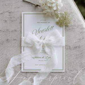 May include: A white wedding invitation with green border, featuring the text "Join us for the wedding of Scarlett Ai". It is tied with a white ribbon and adorned with white flowers and greenery. The invitation is for a wedding in Provence, France.
