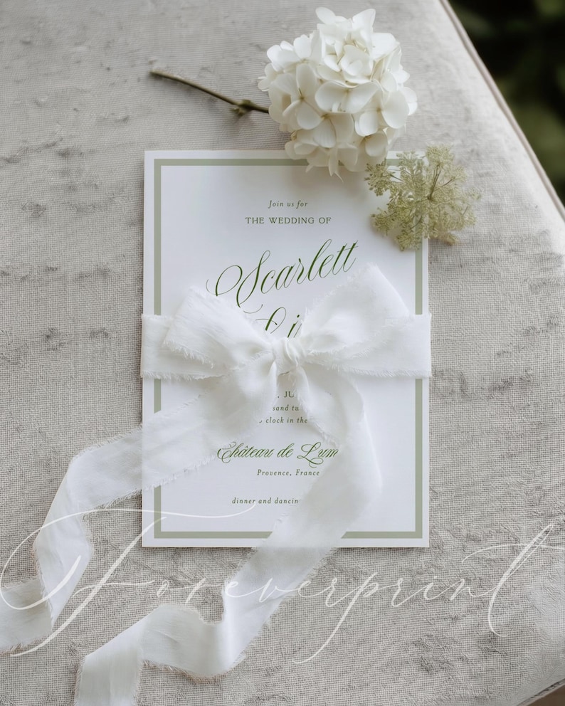 May include: A white wedding invitation with green border, featuring the text "Join us for the wedding of Scarlett Ai". It is tied with a white ribbon and adorned with white flowers and greenery. The invitation is for a wedding in Provence, France.