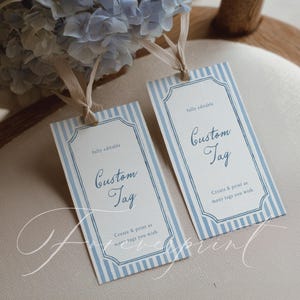 May include: Two rectangular custom tags with blue and white striped borders and the words "Custom Tag" in elegant script. The tags have a cream-colored background and are tied with beige ribbons. The text "Create & print as many tags you wish" is also visible.