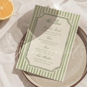 May include: A green and white striped bar menu card with a decorative border sits on a speckled plate. The menu lists signature drinks, wines, beers, and non-alcoholic beverages. A slice of orange and a sprig of wheat are also in the image.