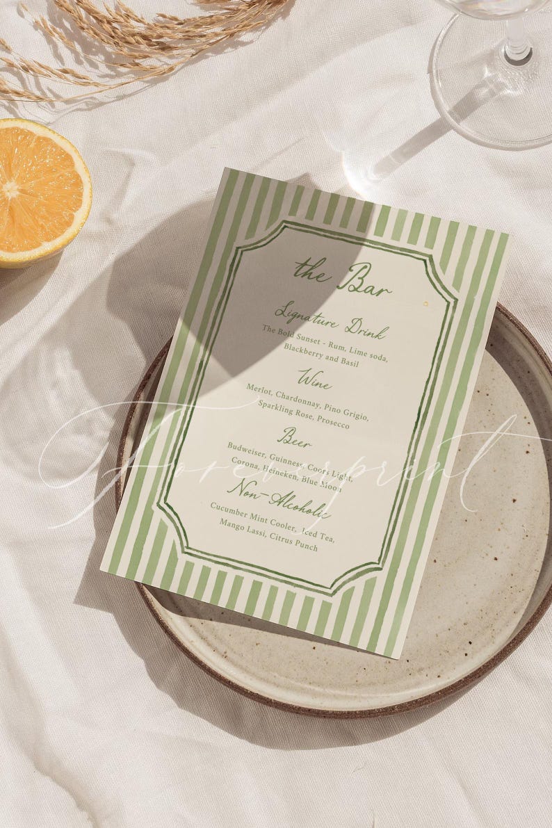 May include: A green and white striped bar menu card with a decorative border sits on a speckled plate. The menu lists signature drinks, wines, beers, and non-alcoholic beverages. A slice of orange and a sprig of wheat are also in the image.