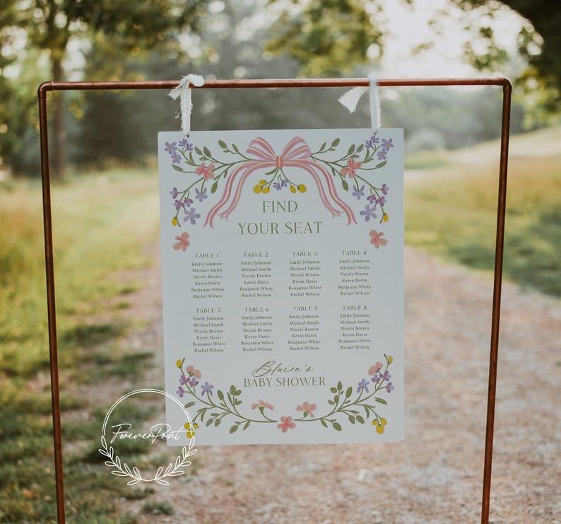 May include: A white seating chart with a floral design and pink ribbon. The chart has the text "Find Your Seat" and lists tables with names for a baby shower. The text "Blaize's Baby Shower" is at the bottom of the chart.