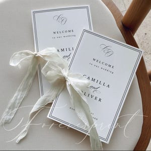 K&ouml;nnte beinhalten: Zwei wei&szlig;e Hochzeits-Willkommenskarten mit schwarzem Text und einem doppelten schwarzen Rand, gebunden mit einem wei&szlig;en Band. Auf den Karten steht "WILLKOMMEN zu unserer Hochzeit" und die Namen "CAMILLA und OLIVER".