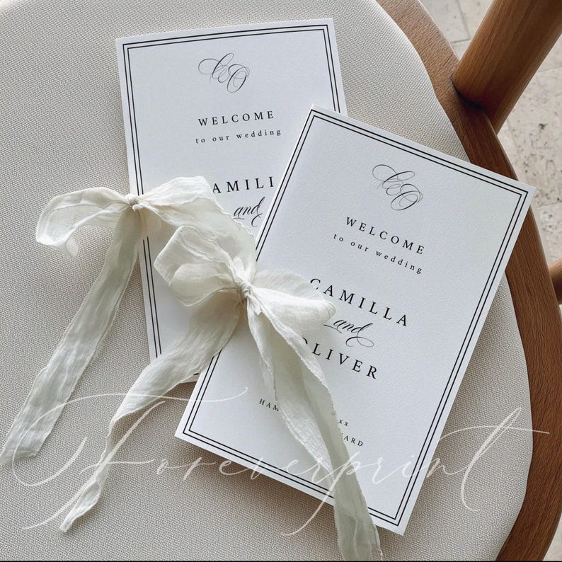 K&ouml;nnte beinhalten: Zwei wei&szlig;e Hochzeits-Willkommenskarten mit schwarzem Text und einem doppelten schwarzen Rand, gebunden mit einem wei&szlig;en Band. Auf den Karten steht "WILLKOMMEN zu unserer Hochzeit" und die Namen "CAMILLA und OLIVER".