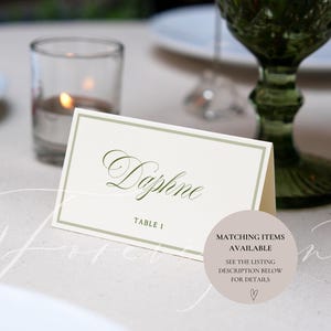 May include: A cream-colored place card with the name "Daphne" in elegant script, framed by a subtle green border. The card is set on a light-colored surface, with a green glass and candle in the background. Table 1 is printed below the name.