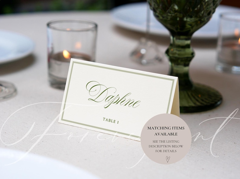 May include: A cream-colored place card with the name "Daphne" in elegant script, framed by a subtle green border. The card is set on a light-colored surface, with a green glass and candle in the background. Table 1 is printed below the name.