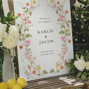 May include: A white wedding sign with a floral border. The sign reads "Welcome to the wedding of Karlie & Jacob, Cleveland, Ohio, 14th August 20xx".