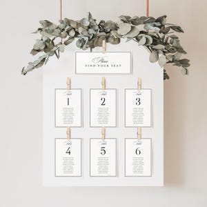 May include: A white seating chart with six table assignments, each held by a wooden clip. The top of the chart reads "Please Find Your Seat." Eucalyptus greenery and copper-colored wire hang above the chart.