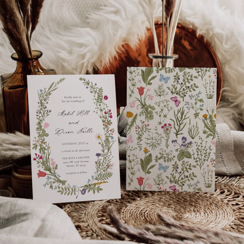 May include: Two wedding invitations. One features a floral wreath with text, including names and date. The other has a floral pattern with butterflies. The invitations are on a textured surface with decorative elements.