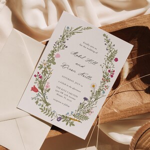 May include: Wedding invitation featuring floral wreath with text. The invitation includes the names "Mobel Hill and Dixen Keulle", date "Saturday, July 14", and venue details. The design incorporates colorful flowers and greenery.