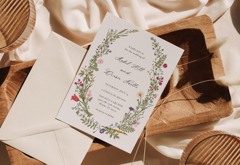 May include: Wedding invitation featuring floral wreath with text. The invitation includes the names "Mobel Hill and Dixen Keulle", date "Saturday, July 14", and venue details. The design incorporates colorful flowers and greenery.