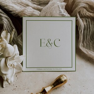 May include: A white card with a green border and the text "E&C" in a script font. Below the text is the phrase "Formal Invitation to Follow". The card is sitting on a beige surface with a white flower and a gold wax seal.