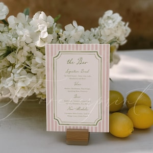 May include: A pink and white striped bar menu with a decorative border, held in a wooden stand. The menu lists signature drinks, wine, beer, and non-alcoholic beverages. Lemons and white flowers are in the background.