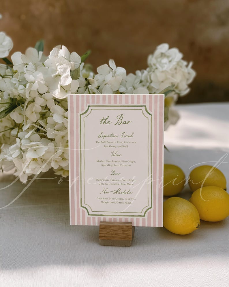 May include: A pink and white striped bar menu with a decorative border, held in a wooden stand. The menu lists signature drinks, wine, beer, and non-alcoholic beverages. Lemons and white flowers are in the background.