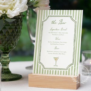May include: A green and white striped bar menu with the text "the Bar" and a list of drinks. The menu is held in a wooden stand. A green glass and white flowers are in the background.