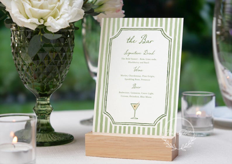 May include: A green and white striped bar menu with the text "the Bar" and a list of drinks. The menu is held in a wooden stand. A green glass and white flowers are in the background.