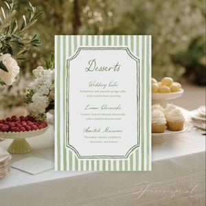 May include: A dessert menu with green and white striped borders lists wedding cake, lemon cheesecake, and assorted macarons. The menu is on a white tablecloth, with desserts and flowers in the background.