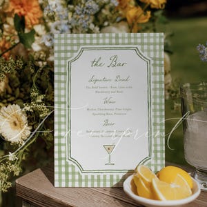 May include: A green and white gingham bar menu with a decorative border. The menu lists signature drinks, wine, and beer options. A small bowl of lemon slices sits in front of the menu, with floral arrangements in the background.