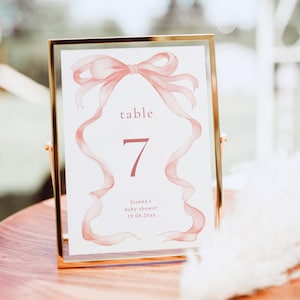 May include: A gold-framed table number card with a pink watercolor ribbon design. The card reads "table 7" and "Sienna's baby shower 19.08.20xx".