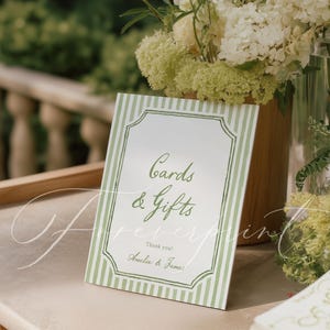 May include: A white sign with green striped borders and the words "Cards & Gifts" in elegant script. The sign also includes "Thank you! Amelia & James." A floral arrangement is in the background.