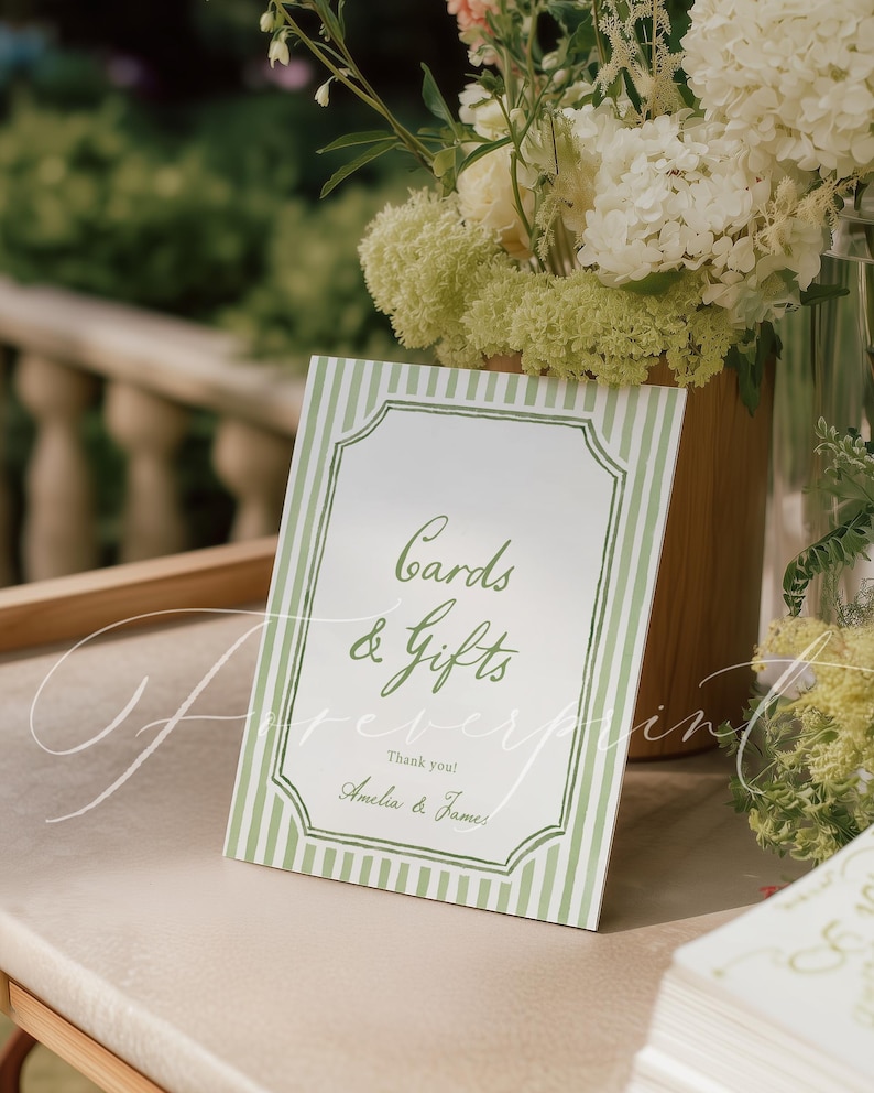 May include: A white sign with green striped borders and the words "Cards & Gifts" in elegant script. The sign also includes "Thank you! Amelia & James." A floral arrangement is in the background.