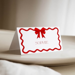 Puede incluir: Una tarjeta de sitio blanca con un lazo rojo y el nombre "SOPHIE" en rojo. La tarjeta tiene un borde rojo ondulado. La tarjeta se muestra en un plato blanco.