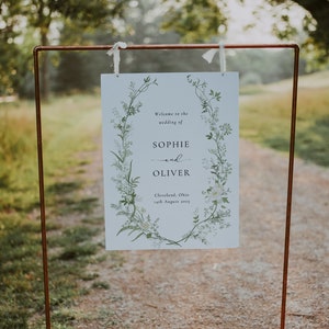 Pode incluir: Um sinal de casamento branco com uma borda floral e o texto "Bem-vindos ao casamento de Sophie e Oliver, Cleveland, Ohio, 14 de agosto de 2023".