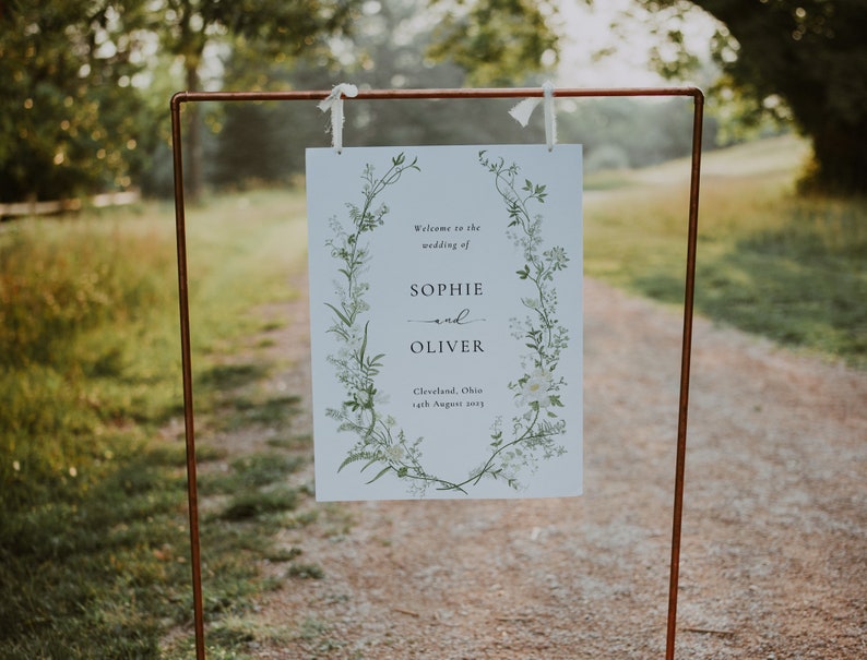 Pode incluir: Um sinal de casamento branco com uma borda floral e o texto "Bem-vindos ao casamento de Sophie e Oliver, Cleveland, Ohio, 14 de agosto de 2023".