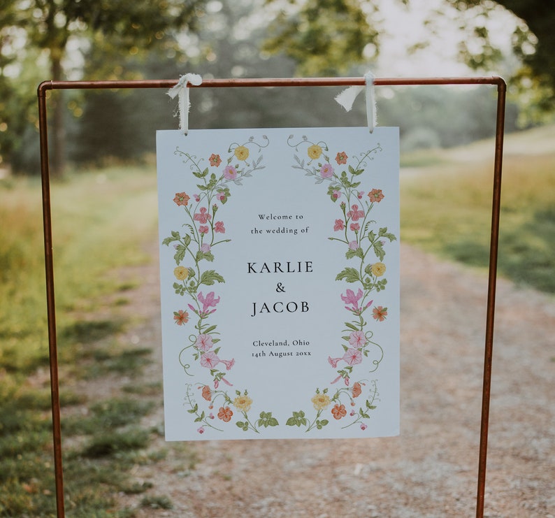 May include: A white wedding sign with a floral border. The sign reads "Welcome to the wedding of Karlie & Jacob, Cleveland, Ohio, 14th August 20XX".