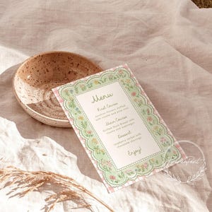 May include: A menu card with a floral border and a pink gingham background. The menu lists a first course of zucchini flowers, a main course of duck breast, and a dessert of raspberry sorbet. A speckled ceramic bowl is also in the image.