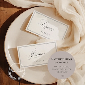 May include: Two elegant place cards with names "Lauren" and "James" in script, each labeled "TABLE 1", rest on a cream-colored plate. A sheer, ivory fabric drapes around the plate. A circular graphic states "MATCHING ITEMS AVAILABLE".