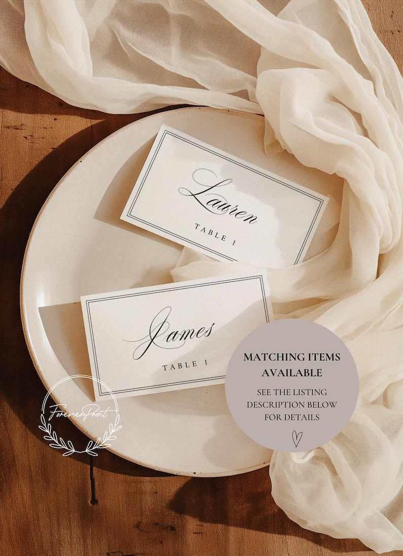 May include: Two elegant place cards with names "Lauren" and "James" in script, each labeled "TABLE 1", rest on a cream-colored plate. A sheer, ivory fabric drapes around the plate. A circular graphic states "MATCHING ITEMS AVAILABLE".