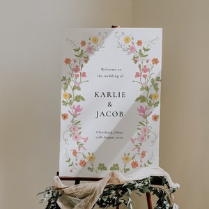 May include: A white wedding sign with a floral border. The sign reads "Welcome to the wedding of Karlie & Jacob, Cleveland, Ohio, 14th August 20xx".