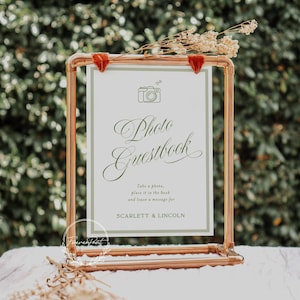 May include: A copper-colored metal frame holds a white sign with the words "Photo Guestbook" in elegant script. The sign also includes a camera graphic and the names Scarlett & Lincoln. Dried flowers and red velvet accents adorn the frame.