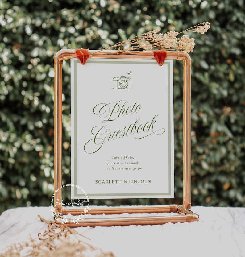May include: A copper-colored metal frame holds a white sign with the words "Photo Guestbook" in elegant script. The sign also includes a camera graphic and the names Scarlett & Lincoln. Dried flowers and red velvet accents adorn the frame.