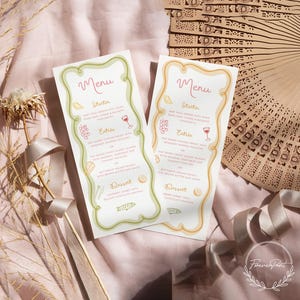 Op de afbeelding: Twee menukaarten met een bloemmotief, met een menu met voorgerechten, hoofdgerechten en dessertopties. De menu's zijn uitgevoerd in een lichtgroene en perzikkleurige kleurstelling. Een decoratieve waaier en lint staan op de achtergrond.
