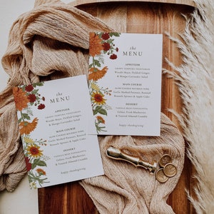 Pu&ograve; includere: Due menu di ringraziamento con disegni floreali. Le carte sono su una superficie di legno con un tovagliolo di stoffa marrone e sigilli di cera dorati. Il menu include antipasti, piatto principale e dessert. Il testo sulle carte recita "the menu" e "happy thanksgiving".