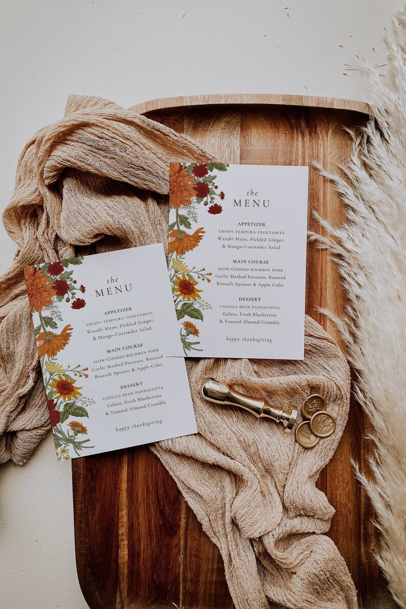 Pu&ograve; includere: Due menu di ringraziamento con disegni floreali. Le carte sono su una superficie di legno con un tovagliolo di stoffa marrone e sigilli di cera dorati. Il menu include antipasti, piatto principale e dessert. Il testo sulle carte recita "the menu" e "happy thanksgiving".
