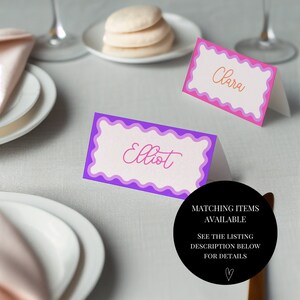 May include: Two place cards with pink and purple scalloped borders. The place cards say "Clara" and "Elliot" in pink script.