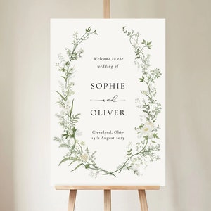 Pode incluir: Um cartaz de boas-vindas de casamento branco com um design de coroa de flores. O cartaz diz "Bem-vindos ao casamento de Sophie e Oliver, Cleveland, Ohio, 14 de agosto de 2023."